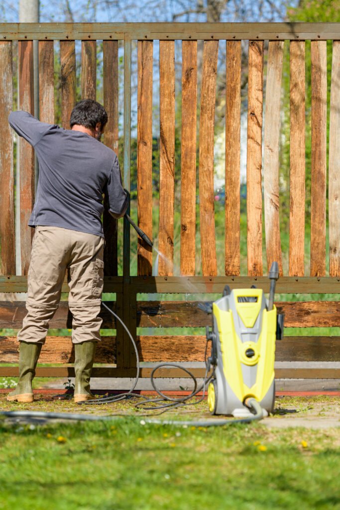 mid adult man cleaning a wooden gate with a power washer. high water pressure cleaner used to diy repair garden gate.